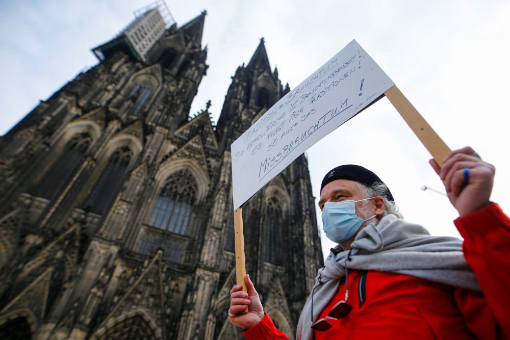 An older white man wearing a surgical mask carries a sign in the shadow of a cathedral