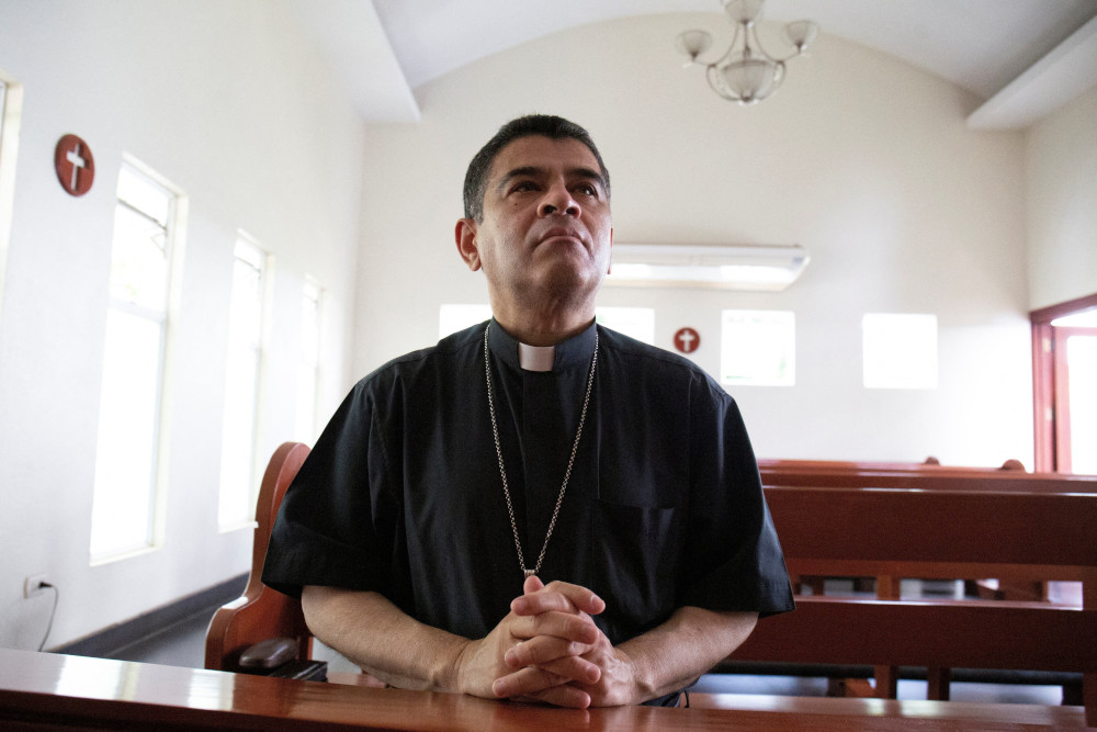 A light brown skinned man wearing a clerical shirt and a pectoral cross folds his hands in prayer