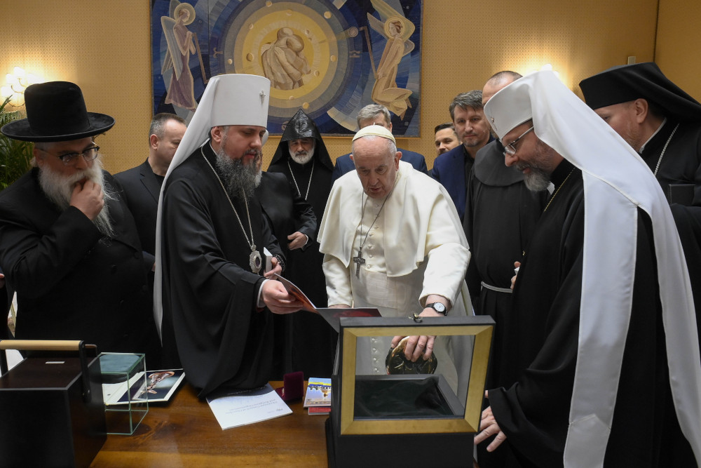 Pope Francis looks inside an open box with a crowd of older men in religious garb standing around him