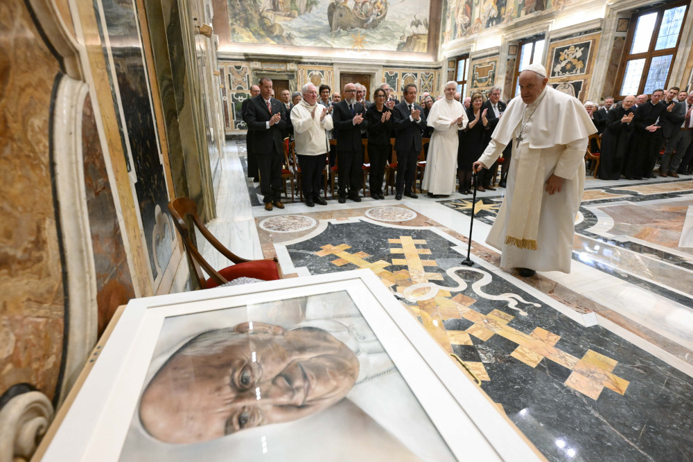 Pope Francis stands with a cane and looks at a portrait of him that is much bigger than him with an audience in the background