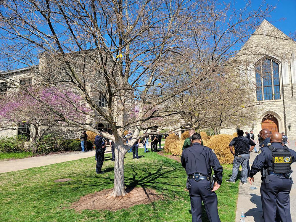 Police officers stand outside next to a tree and a building with a large window and peaked roof