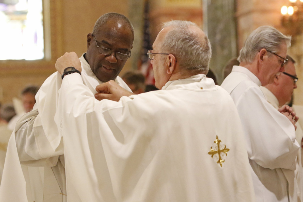 An older white man wearing glasses and a white chasuble adjusts the vestments of a bald Black man