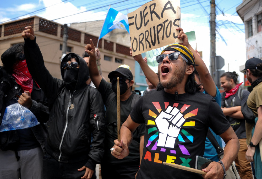 A group of people march in the street. Some wear masks. One holds a sign saying "Fuera Corruptos" high.