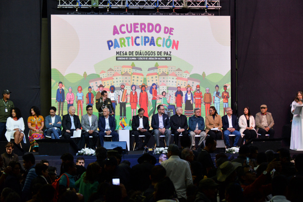 Many people sit in a line under a screen with the words "Acuerdo de Participación" and colorfully illustrated images of people