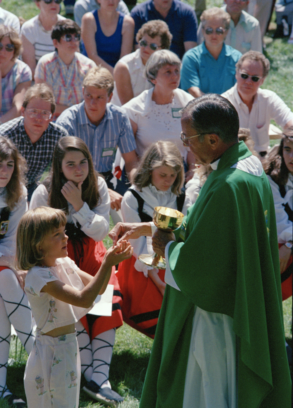 A man in green vestments distributes Communion to a group of white people outside