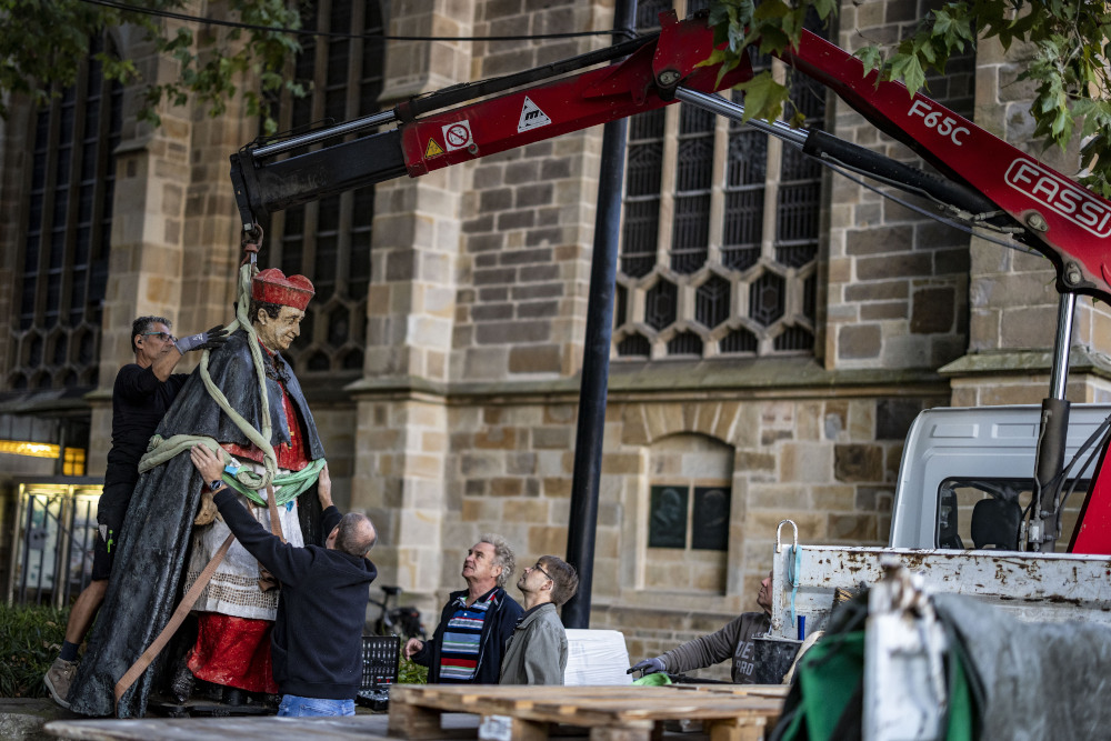 A statue of a man wearing a red zucchetto is lifted by a red crane while men steady the statue and others look on