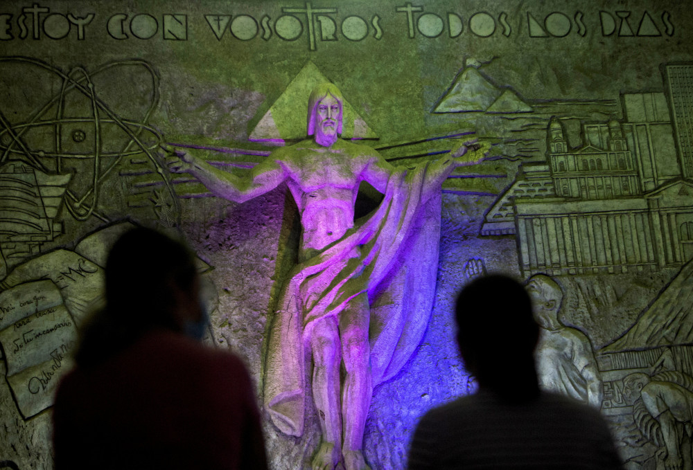 The shadows of two people are visible before a stone statue of Jesus that stands in front of the words, "Estoy con vosotros todos los dias."