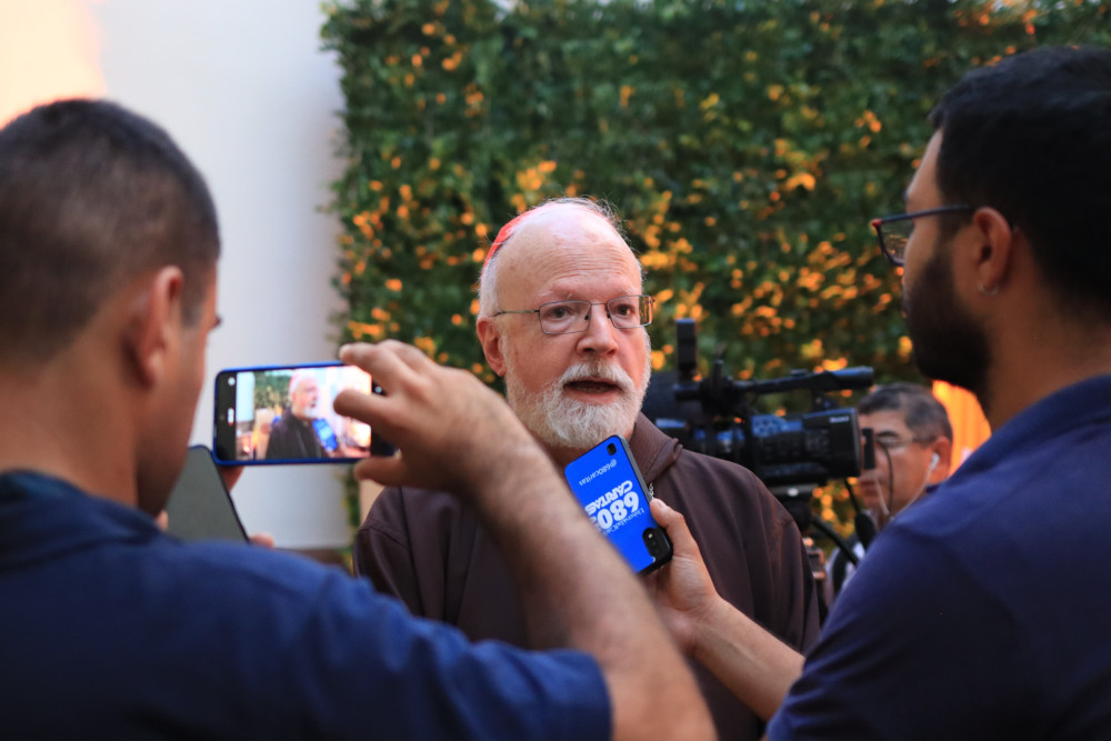 Two people hold phones towards an older white man wearing a brown robe and a red zucchetto