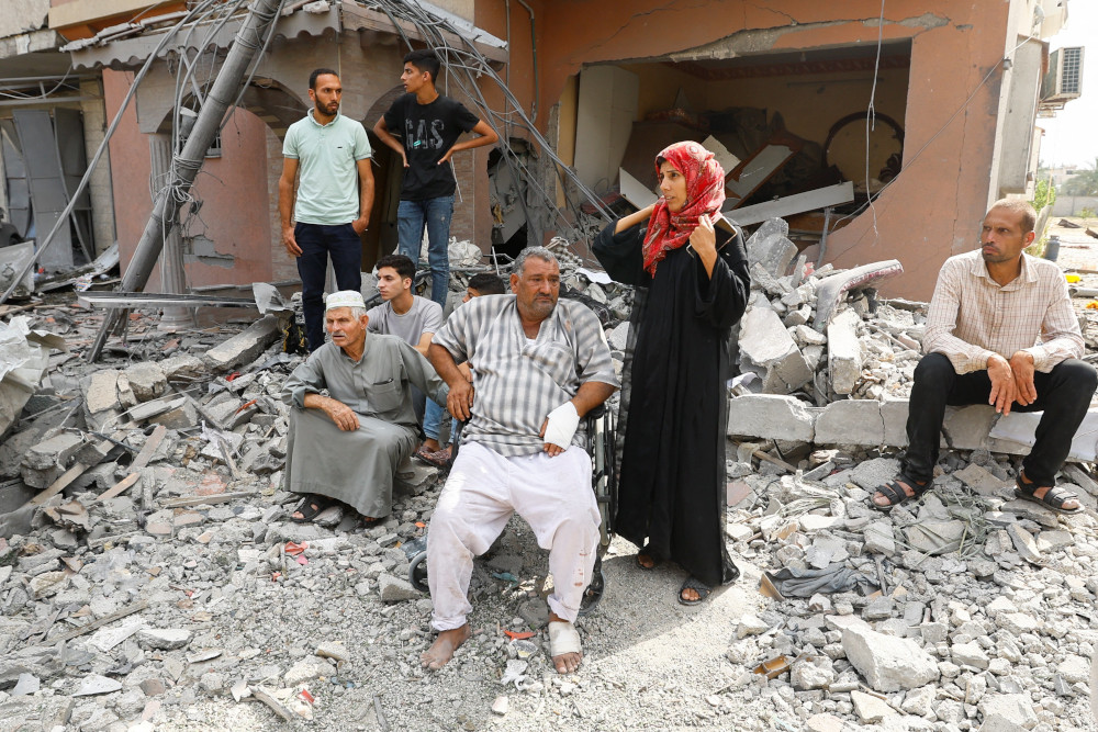 Men and a hijabi woman sit and stand around the rubble of a destroyed building