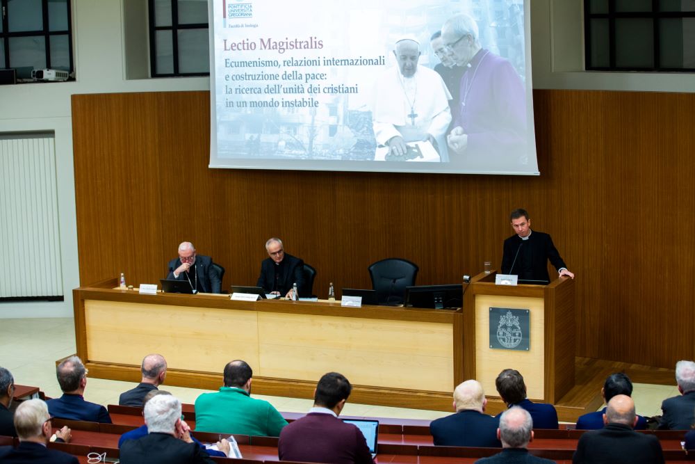 A man stands at a lectern, with two men sitting nearby and a screen projecting the title of the event behind them. An audience watches. 