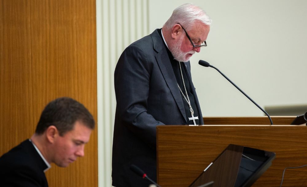 Bishop speaks at microphone. Another cleric listens.