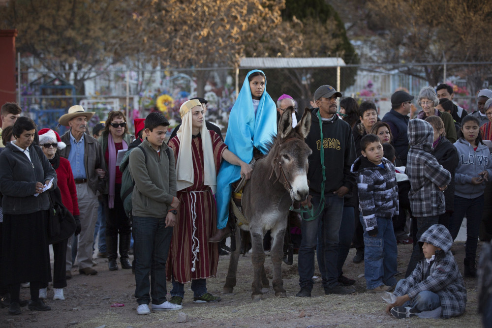 A crowd stands around a girl wearing a blue nativity costume riding on a donkey. A boy also dressed in nativity clothes stands beside her.