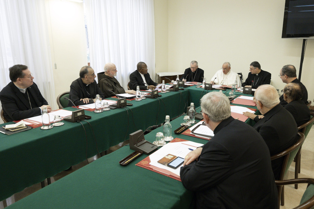 Men wearing black and clerical collars sit around green tables in a U shape with Pope Francis at the head.