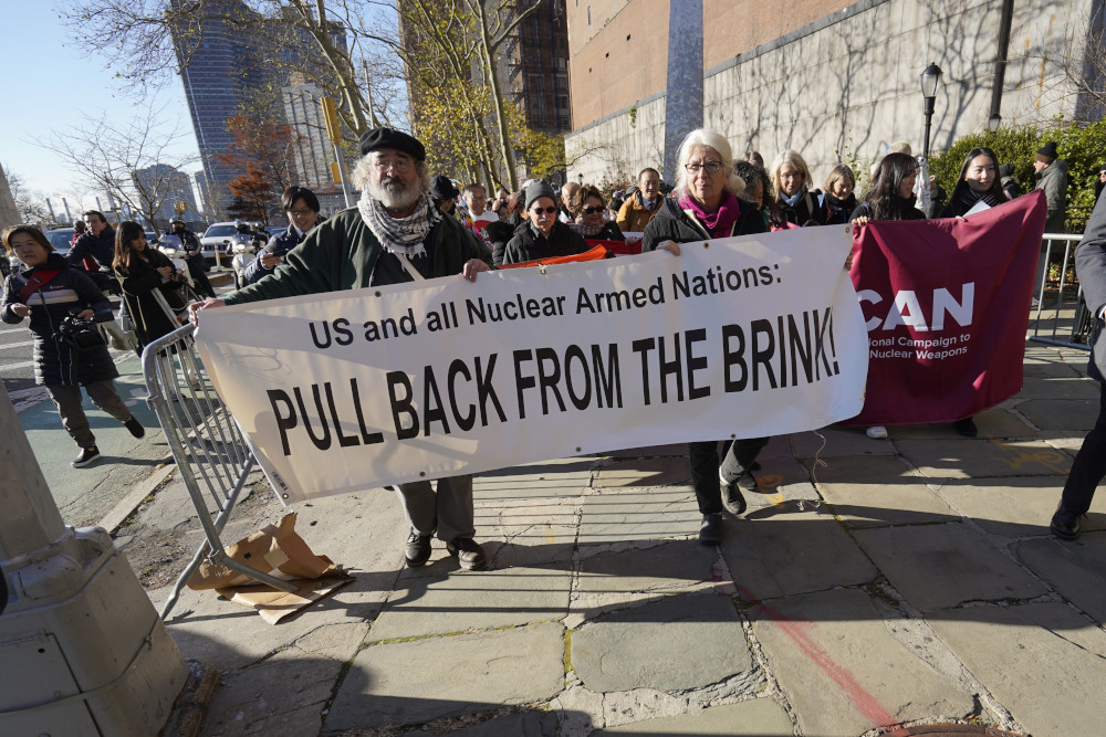 Two people hold a banner that says "U.S. and Nuclear Armed Nations:  Pull Back From The Brink" while walking in front of a crowd