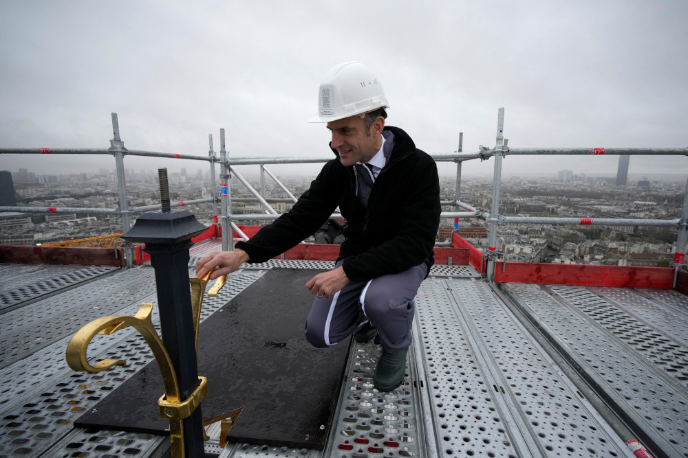 A white man wearing a white hardhat crouches on a metal platform in the sky and touches the gold top of a spire