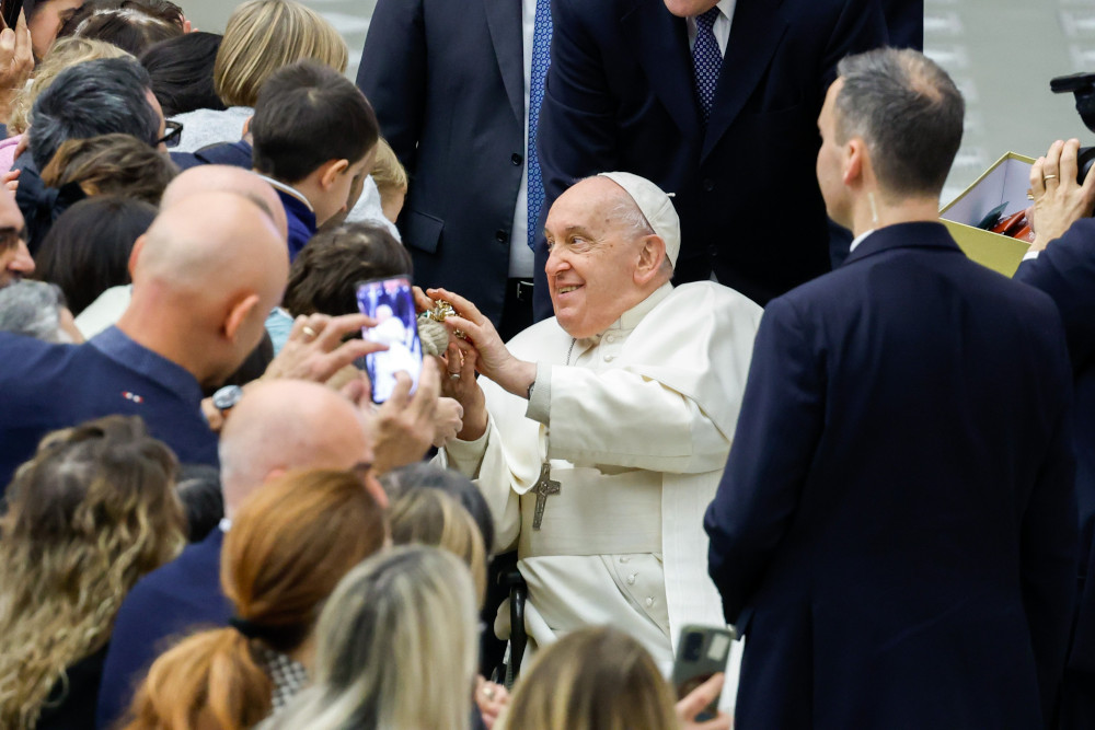 Pope Francis sits in a wheelchair rolling past a crowd of people, turning to the side to rest his hands on an object.