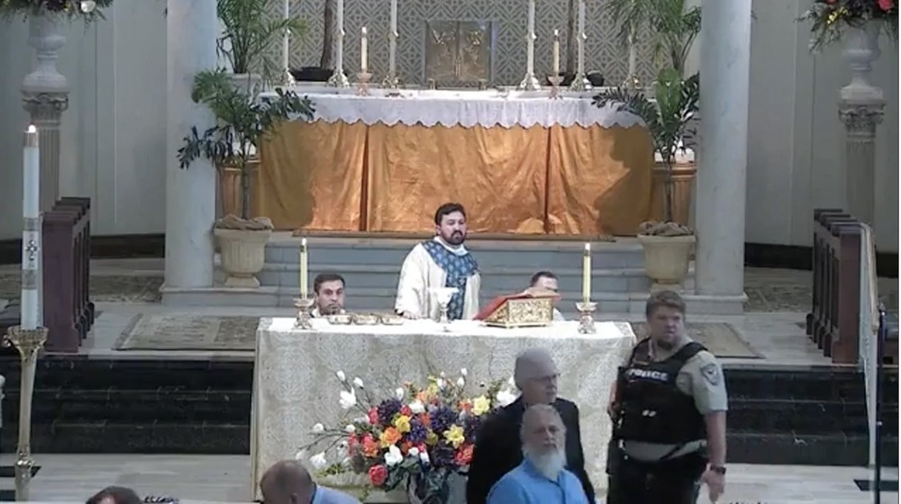 Priest stands at altar while acolytes kneel and police officer scans crowd. 