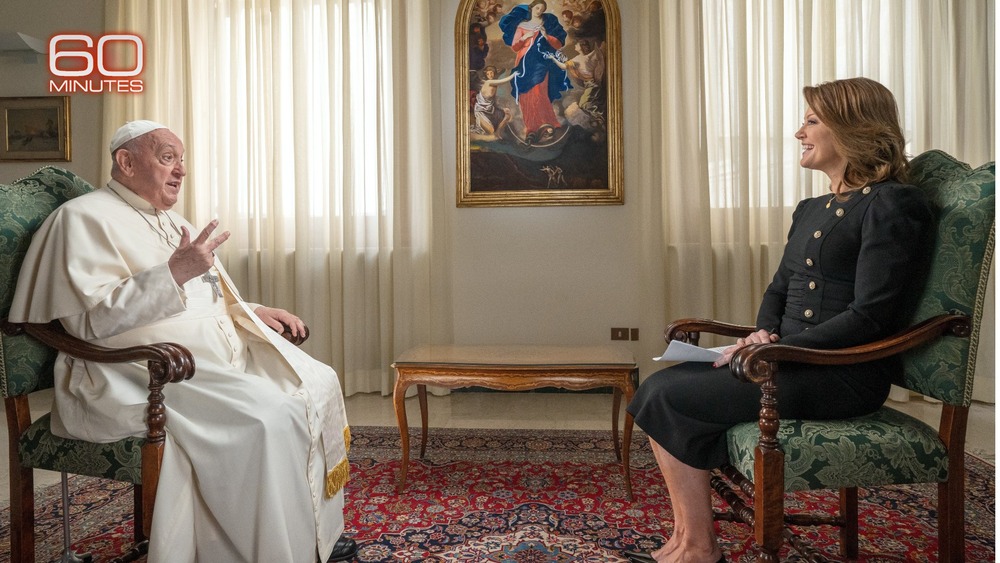 Pope Francis and Norah O'Donnell seated opposite one another, with painting and draped windows in background. 