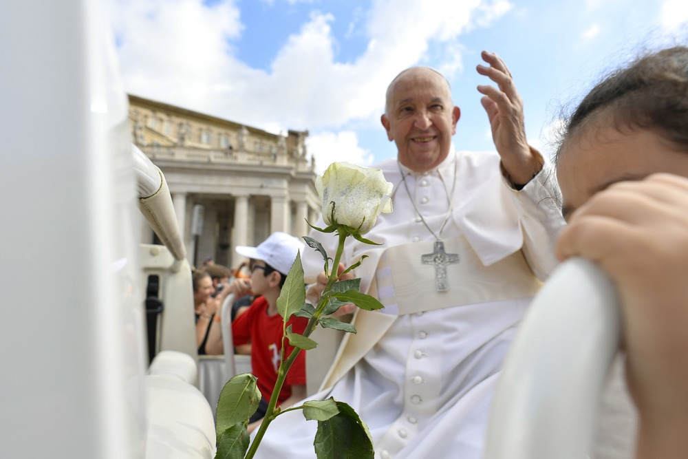Pope Francis, seated, framed by faces and a white rose, as he smiles and raises his hand in blessing. 