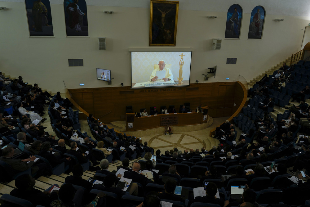 Pope Francis seen on screen in front of large seated assembly. 
