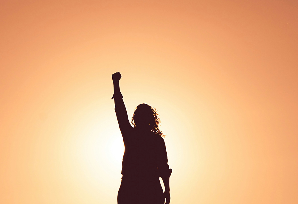 A woman raises a clenched fist into the air. She is silhouetted against a sunlit sky. (Unsplash/Miguel Bruna)
