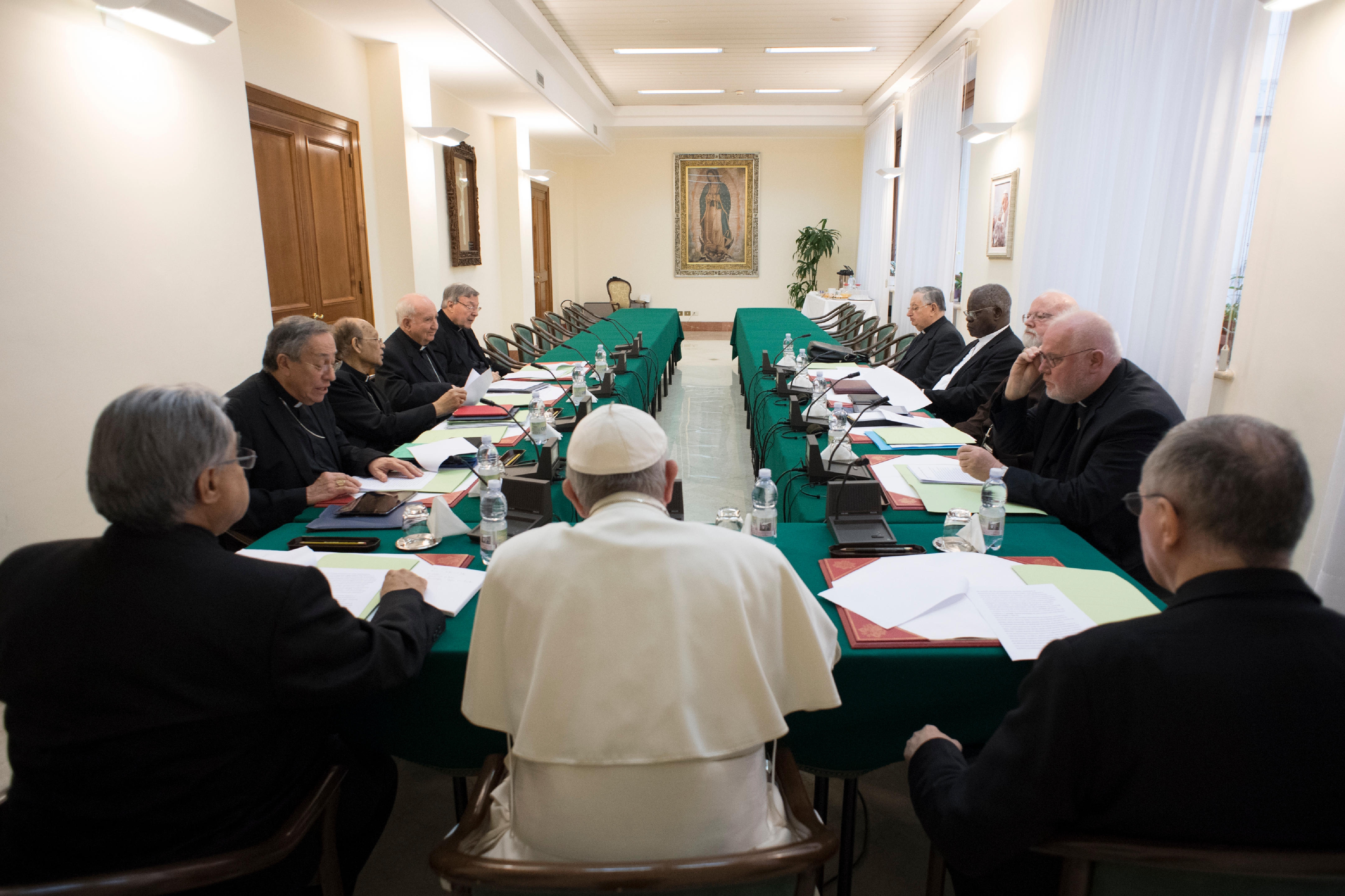 Pope, seated and pictured from behind, surrounded by Cardinals at large U-shaped table, covered in green; Our Lady of Guadalupe hangs on far wall of room. 