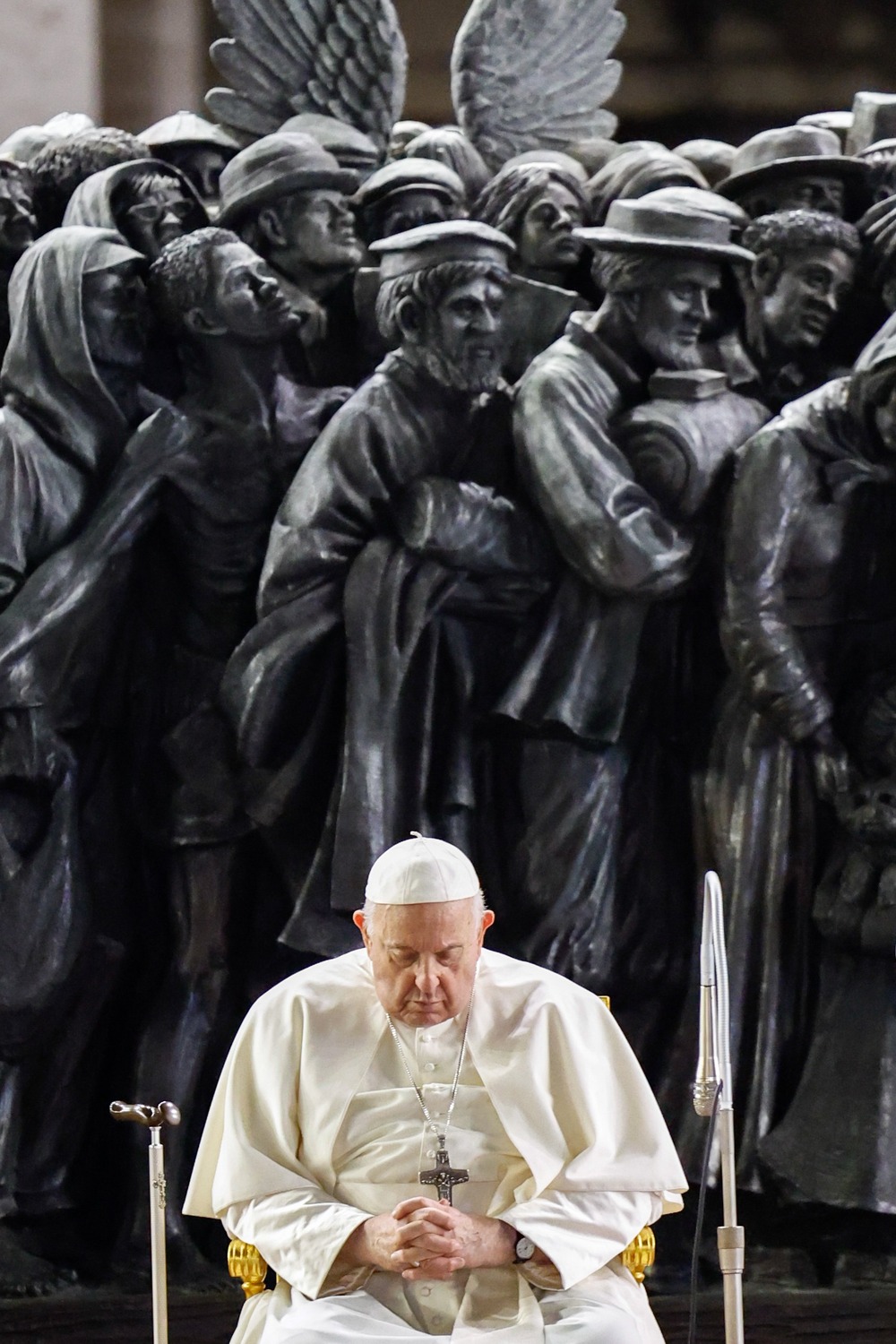 Pope Francis, seated, bows head in prayer, with bronze procession of migrants directly behind. 