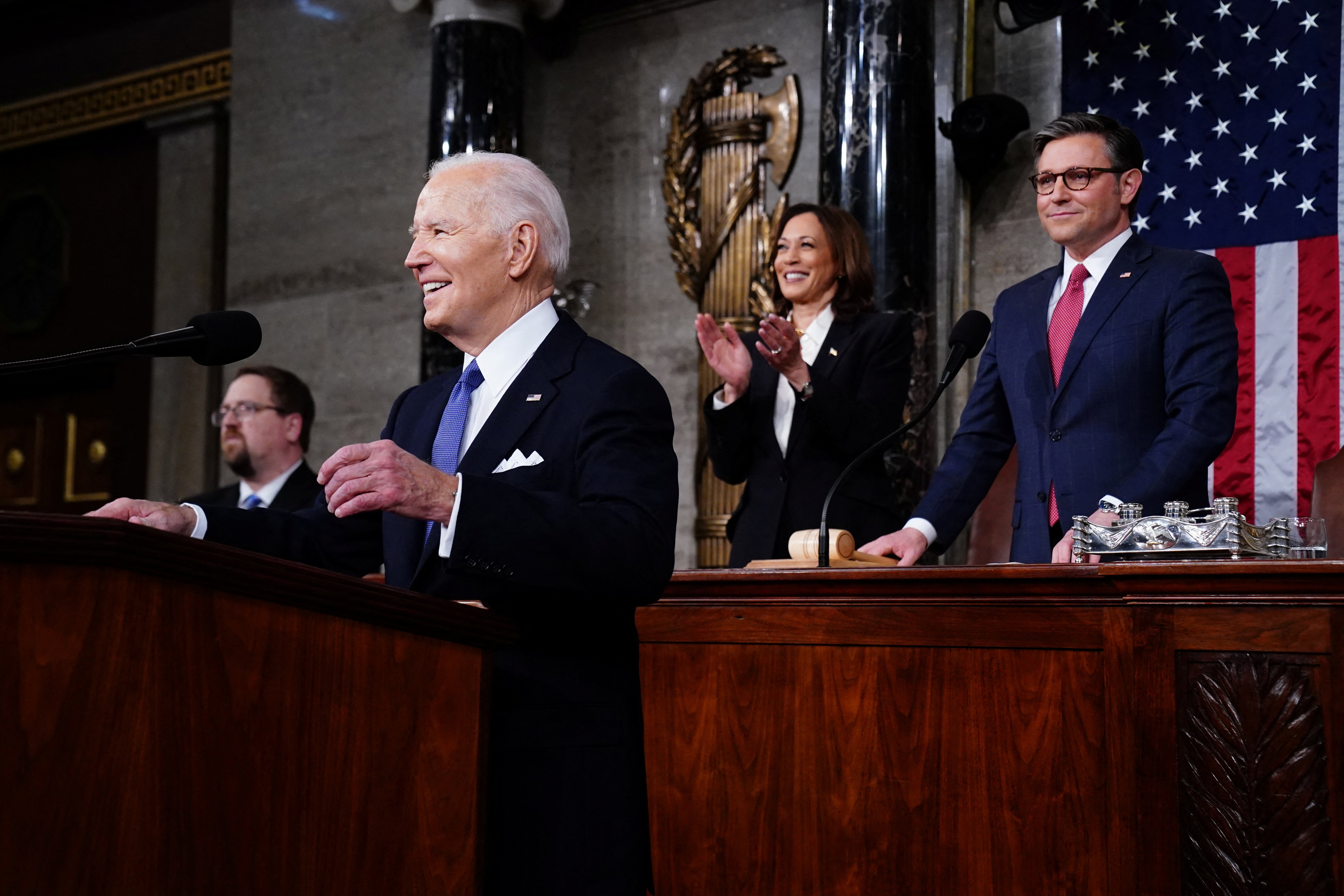 President Biden, smiling, stands at lectern, Vice President Harris and Speaker Johnson stand behind. 