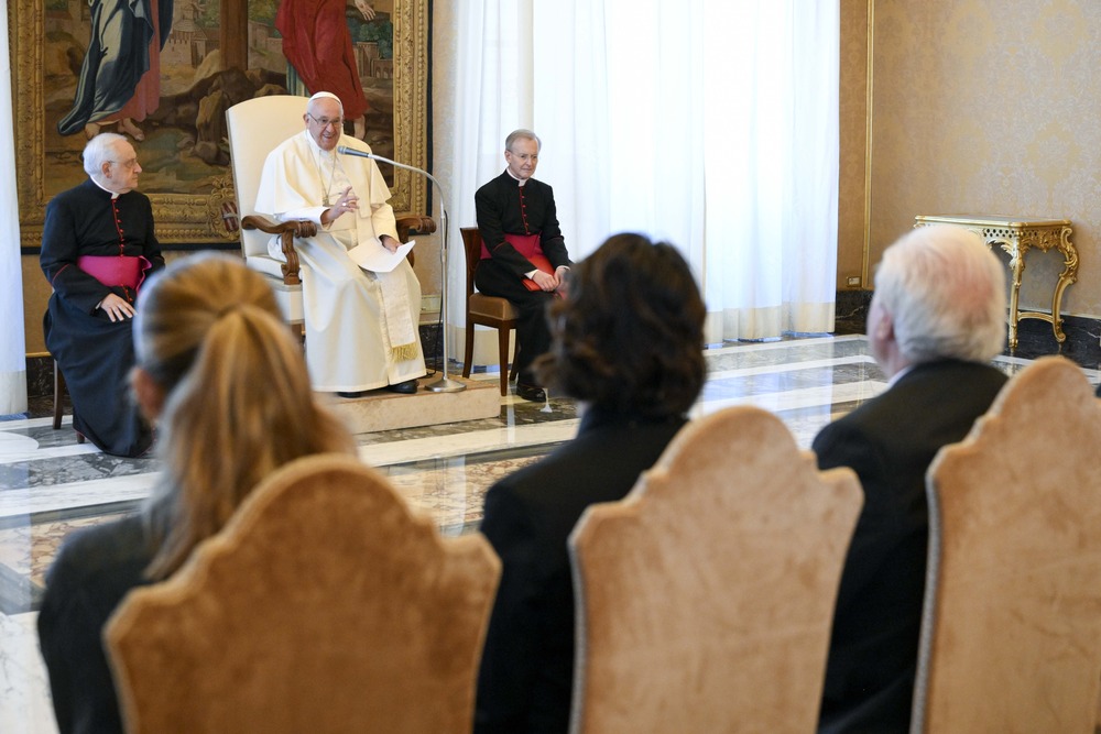 Pope Francis seated, flanked by Cardinals, people sit facing him, the back of their heads to the camera. 