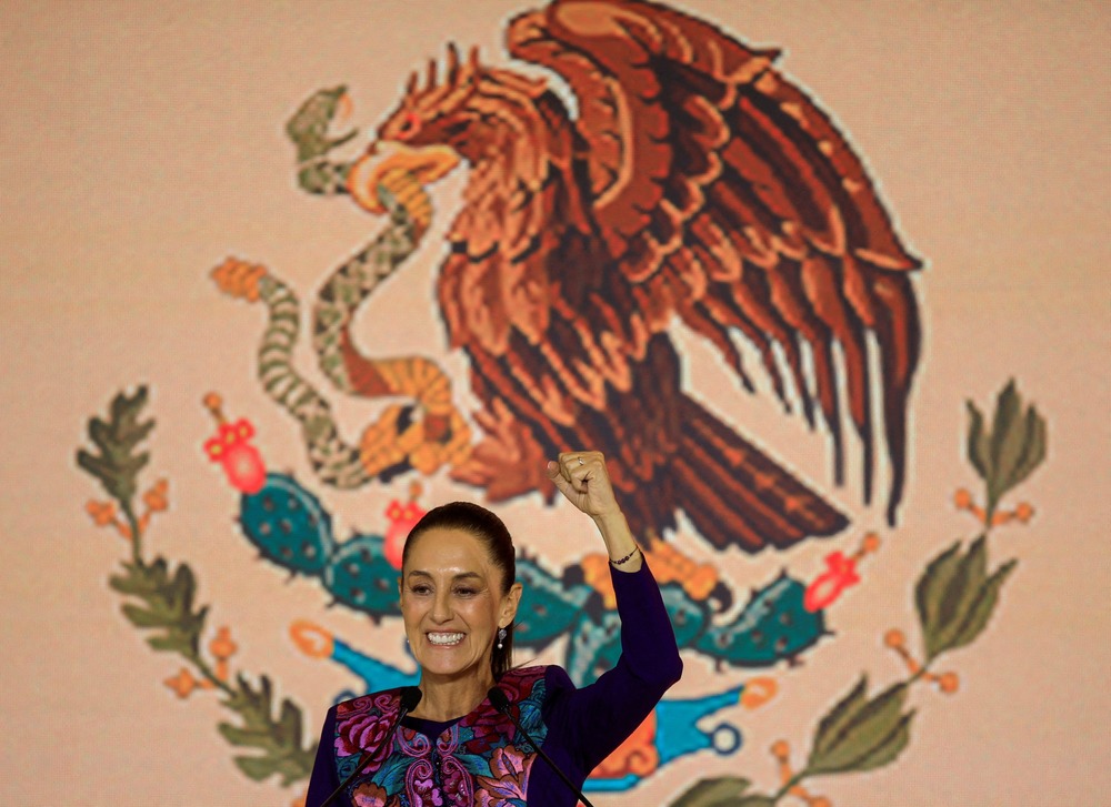 Scheinbaum smiles and raises fist in victory in front of banner bearing Mexico's eagle emblem.