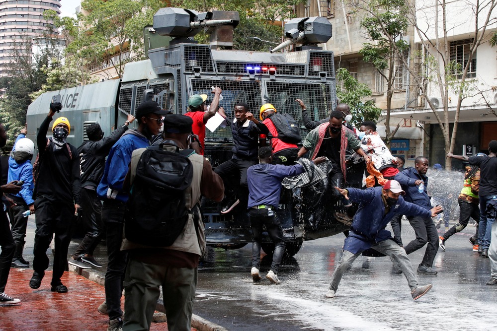 People jump in front of and on large, armored vehicle in street. 