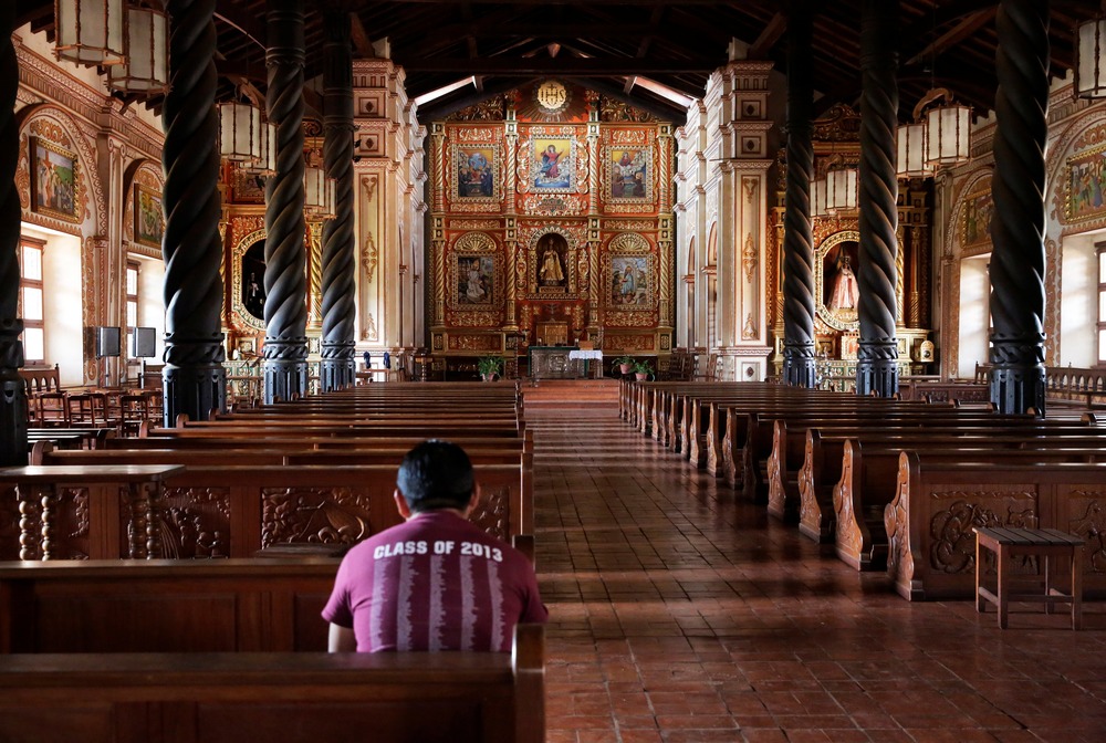 Man pictured from behind, seated in rear pew; aisle, columned nave, and elaborate altar of cathedral visible. 