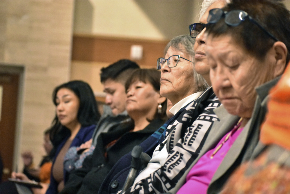 A long line of seated people are shown from the side, their faces somber. 