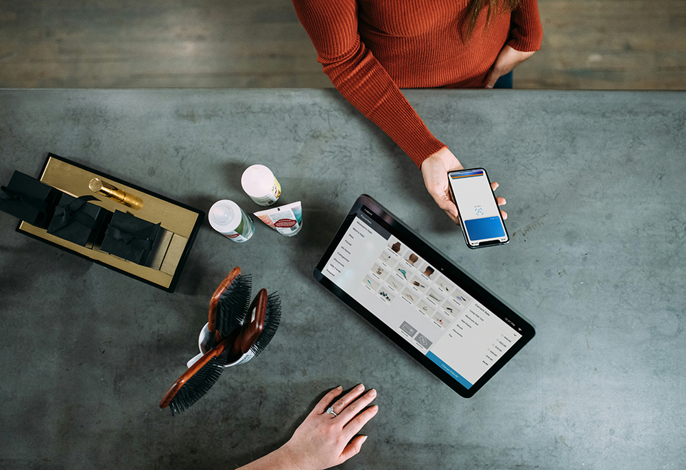 In an overhead photo illustration, a customer uses a smartphone to make a transaction at a salon, while another person uses transaction software on a tablet. (Unsplash/Blake Wisz)