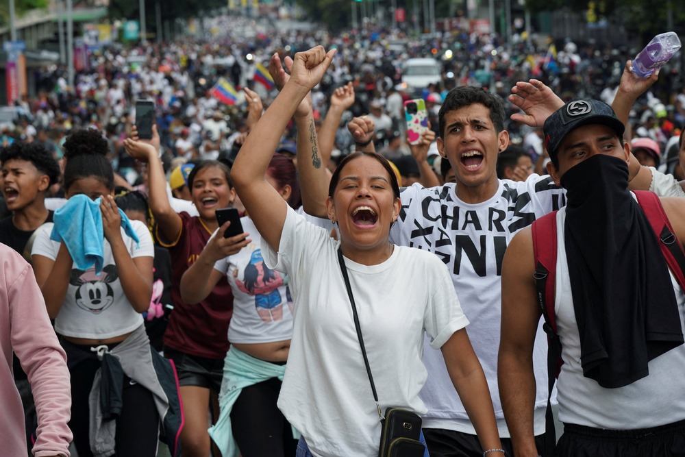 Woman in white shirt shouts chant while raising fist; behind hundreds of white-clothed people assemble and march. 