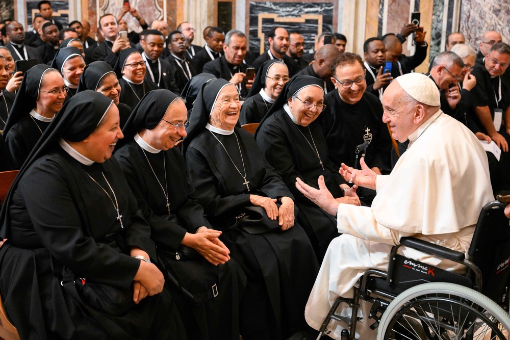 Pope, in wheelchair, sits very near group of habited sisters who all laugh in response to pope's comment. 