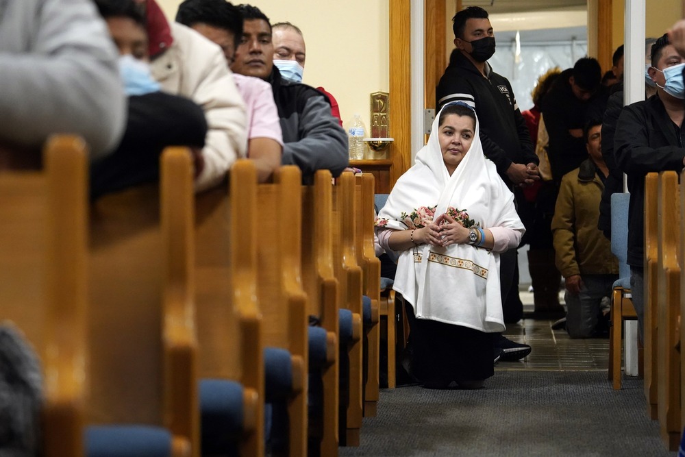 Woman wears white mantle and kneels in church aisle; others kneel in pews. 