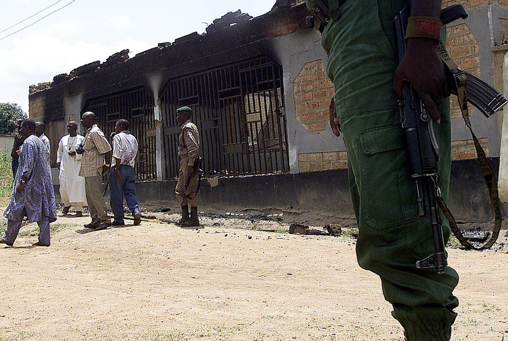 Armed police stand outside burnt shell of building; civilians come and go. 