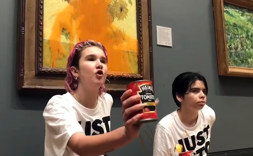 Two young people wearing matching t-shirts, stand in front of painting; one holds can of soup, whose contents are splattered across painting. 