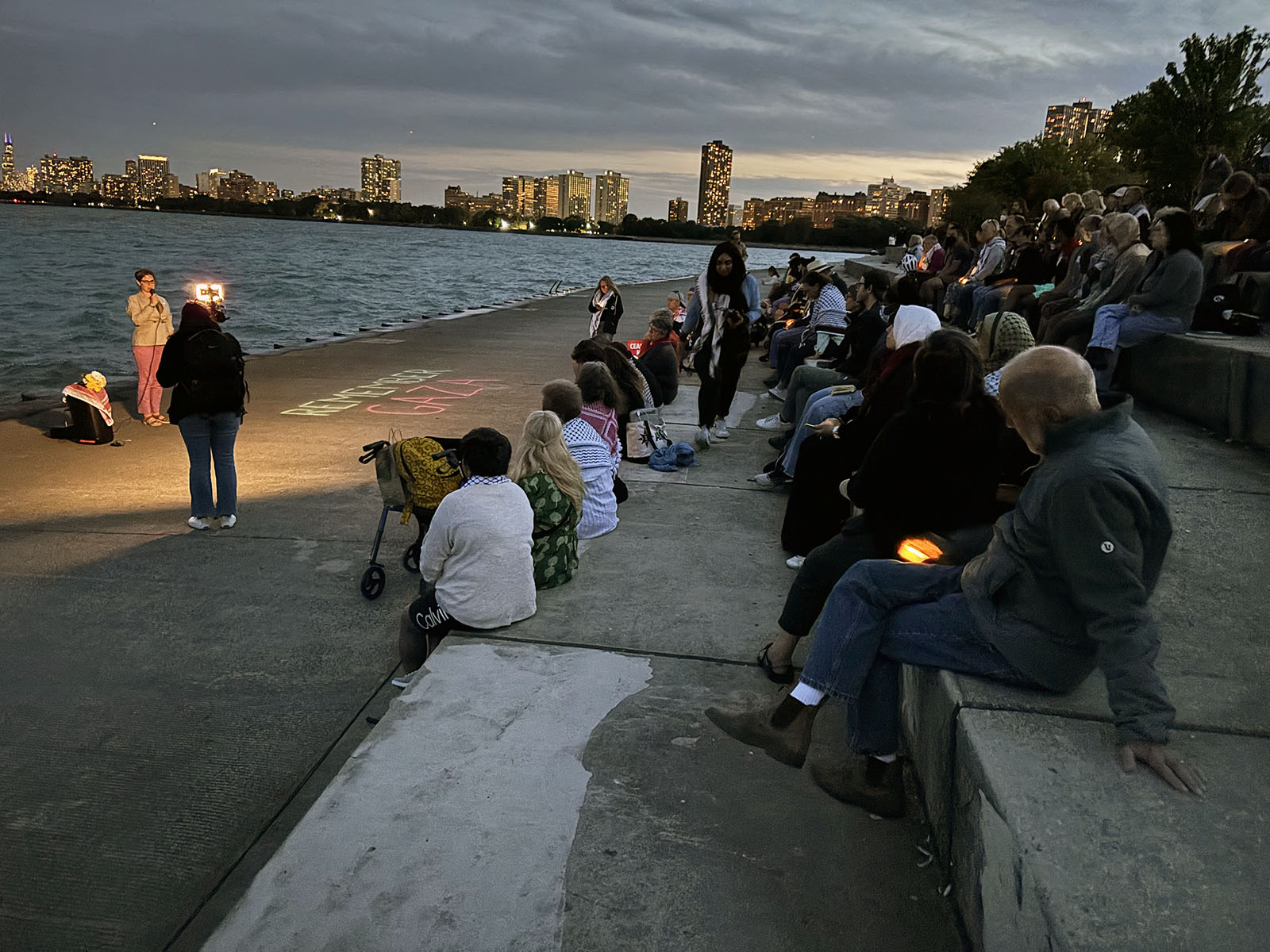 Chicago skyline at dusk in background; people sit gathered around speakers at edge of Lake Michigan. 