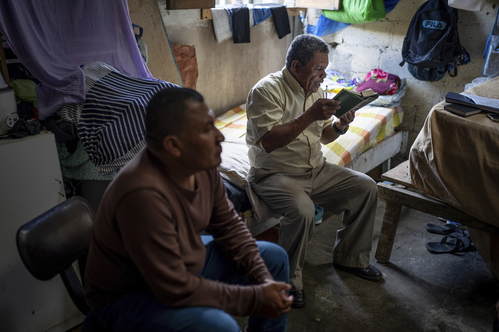 Alvicio sits on a twin bed in a cinderblock room reading from bible, Ocampo sits to his right on a chair.