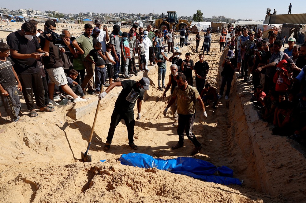 People line up along a ditch, standing in the ditch are the diggers, and wrapped in a blue tarp is a corpse for burial.