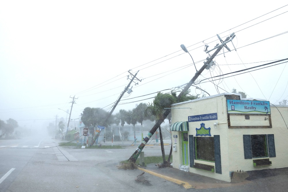 Windswept, rainy view of utility poles fallen; one leans on small building.