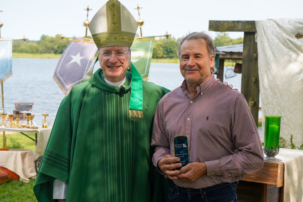 The bishop, vested for ordinary time and wearing mitre, stands with his arm around Kremer who holds his award plaque.