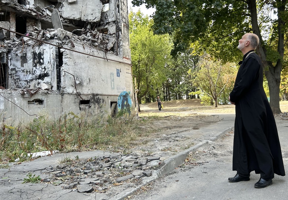 The archbishop wears plain black cassock, and stands back regarding ruins of building.