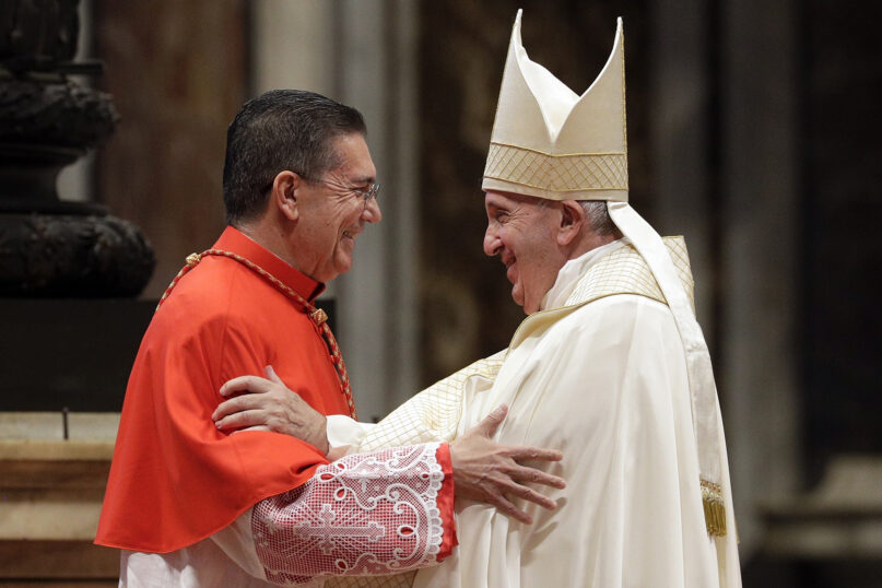 The cardinal embraces the pope, who is vested for Mass in white, wearing mitre.