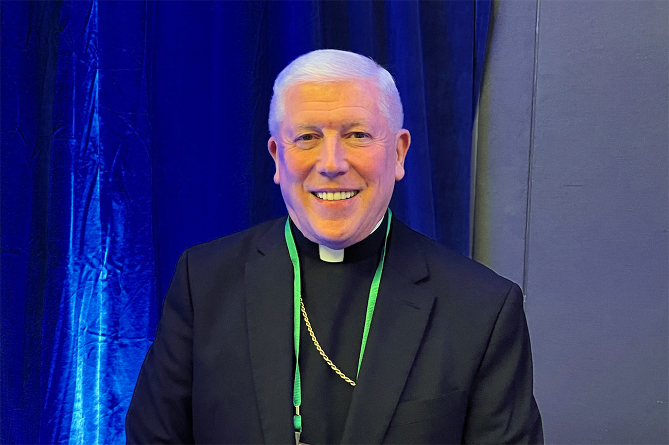 The bishop wears plain black clerics and smiles against a blue backdrop.