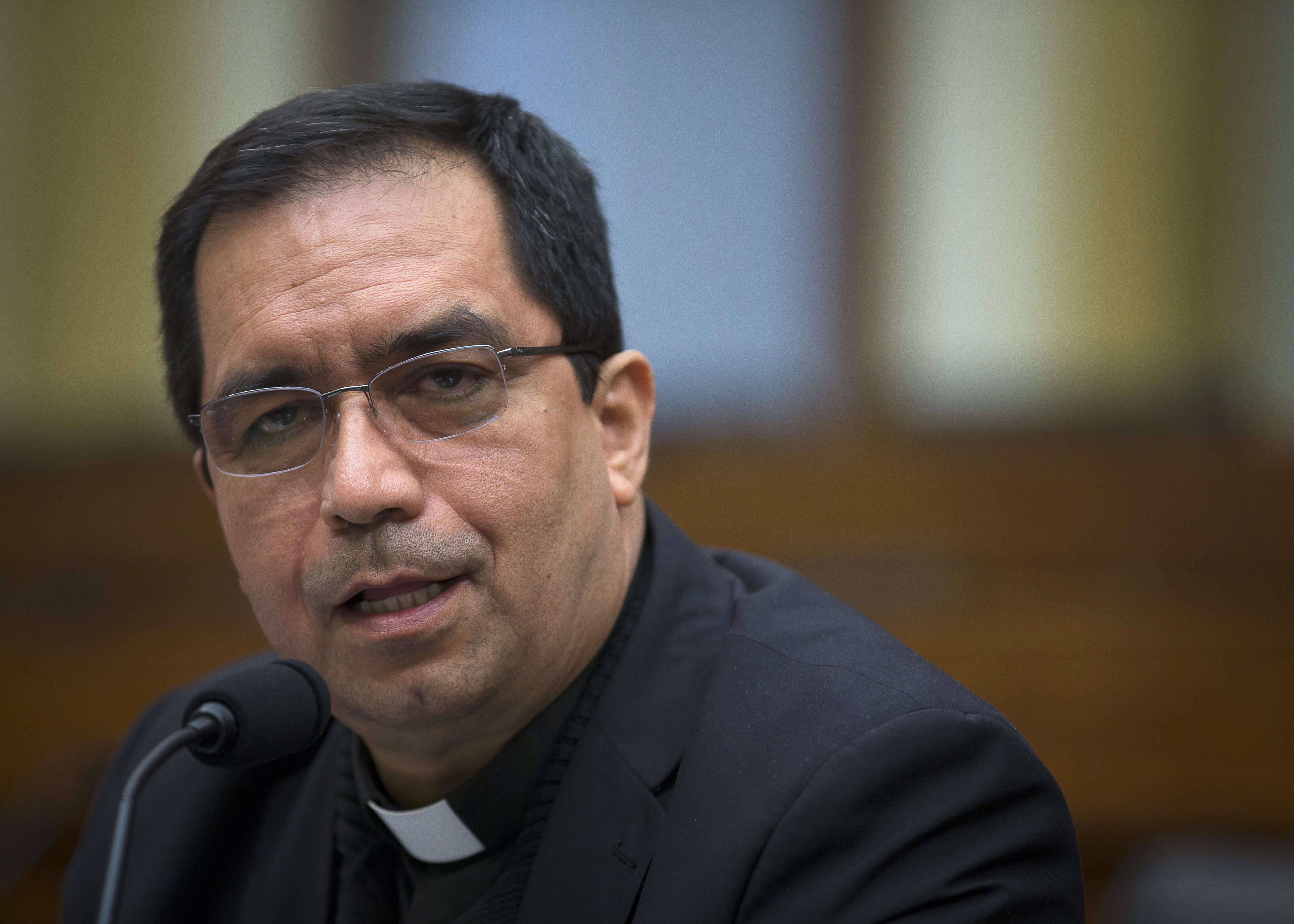 Escobar sits at table, speaking into microphone. He wears plain black clerics.