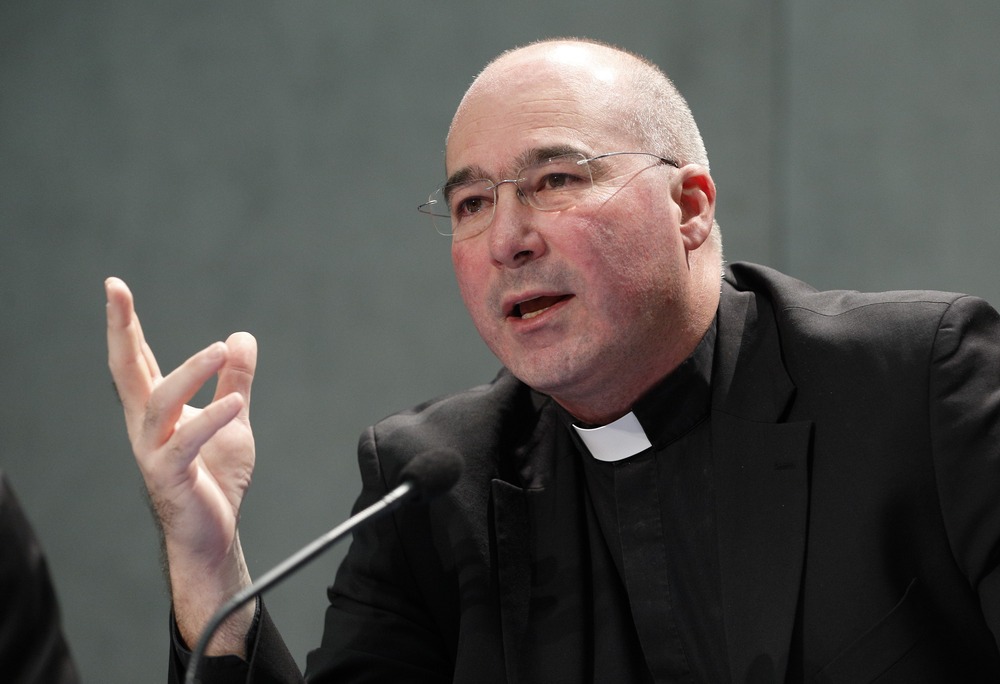 The priest wears black clerics and speaks and gestures into microphone.