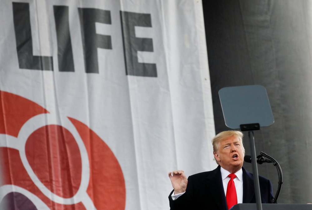 Trump stands at podium in front of large banner gesturing while talking.